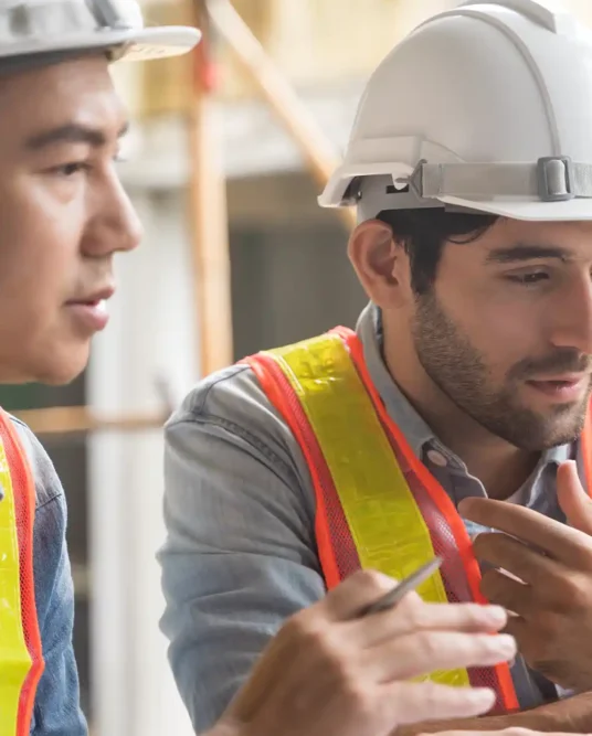 Two field engineers in hard hats review plans on a laptop during a site walk, coordinating IT cabling and electrical work.